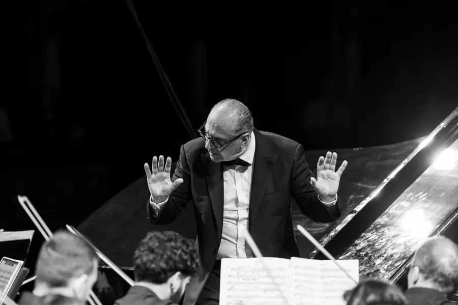 Bianco e nero: in frac e papillon, entrambe le mani sollevate a fermare il suono, la cassa del pianoforte alle spalle e gli spartiti dei professori in primo piano