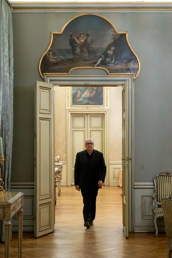 Figura in abito scuro che avanza attraverso una porta a due battenti di un palazzo antico, sotto una sovrapporta dipinta, in un'infilata di sale
