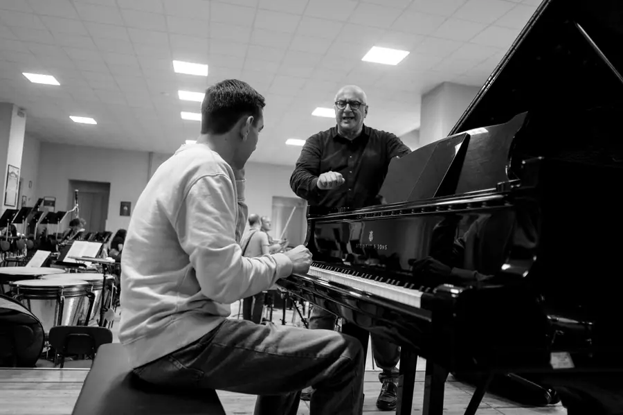 Fotografia in bianco e nero: uomo calvo con occhiali in piedi accanto a un pianoforte a coda aperto, mentre parla e gesticola verso un giovane pianista seduto di spalle; sullo sfondo una sala prove con leggii e timpani