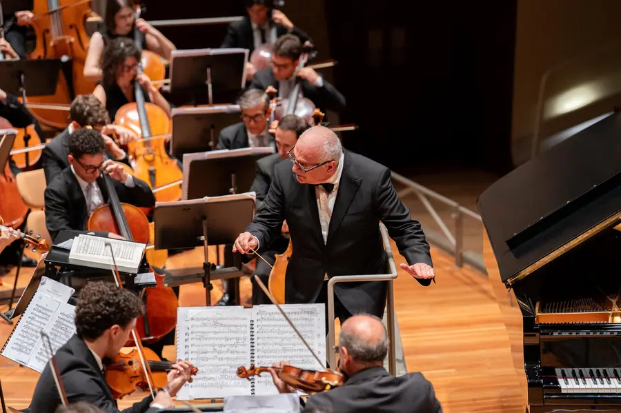 Il direttore in frac di tre quarti mentre dirige, bacchetta alzata sul leggio, violoncelli a sinistra e il coperchio di un pianoforte a coda a destra