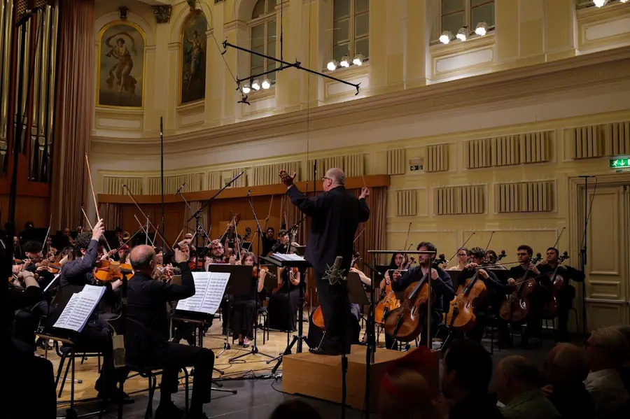Direttore su una pedana di legno con entrambe le braccia sollevate, orchestra d'archi e violoncelli intorno, sala chiara con pannelli e dipinti alle pareti e luce calda.