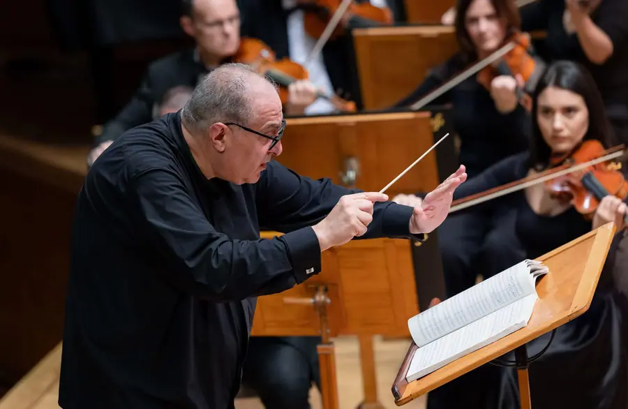 Il direttore proteso in avanti con la bacchetta nella destra e la sinistra aperta, orchestra e leggii alle spalle, partitura aperta sul leggio in primo piano