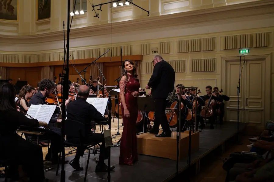 Soprano in abito rosso lungo sul bordo del palco accanto al direttore, che si e' voltato verso di lei; l'orchestra suona alle loro spalle.