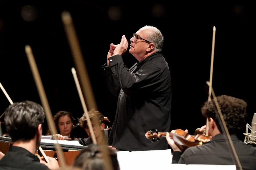 Direttore ripreso di tre quarti con le mani unite davanti al viso, orchestra in primo piano sfocata