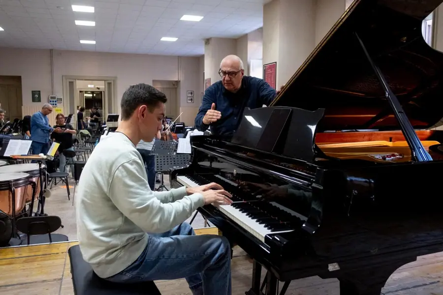 In sala prove, appoggiato al coperchio del pianoforte a coda, parla con un giovane solista seduto alla tastiera mentre l'orchestra sistema gli strumenti sullo sfondo