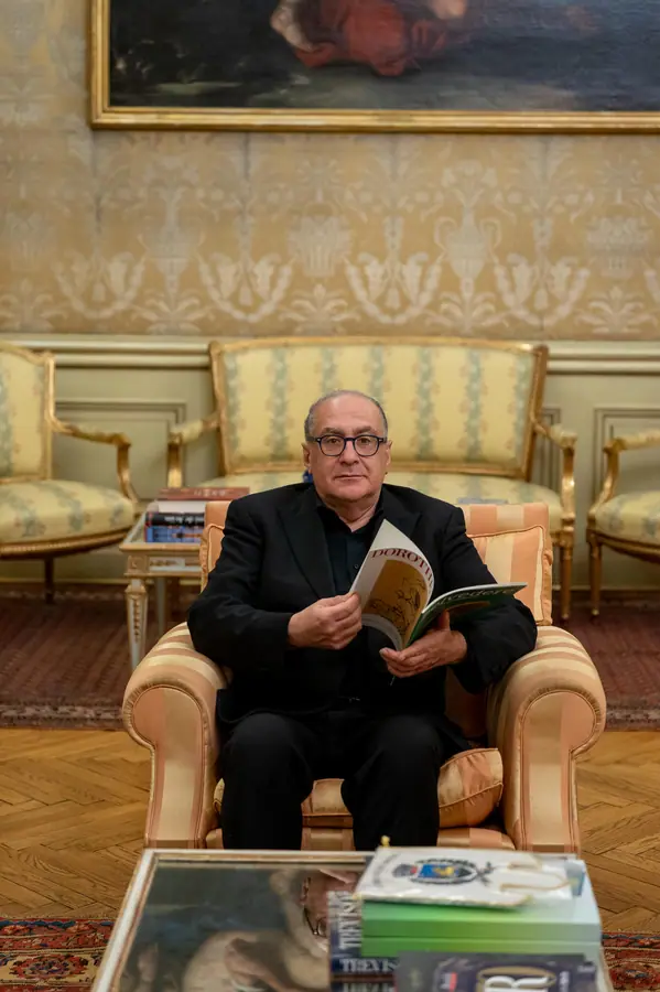 Uomo con occhiali seduto in una poltrona dorata, tiene aperta una rivista; alle spalle un divano damascato oro e un grande dipinto