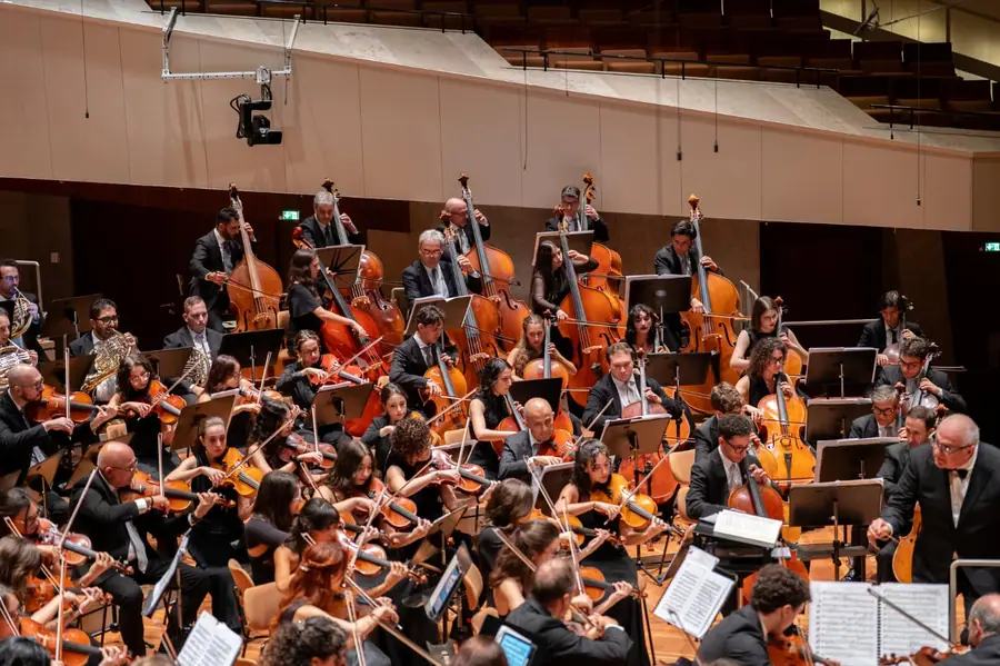 Veduta d'insieme dell'orchestra al completo su un palco moderno in legno chiaro, con i contrabbassi in fondo; a destra, di spalle, la figura del direttore