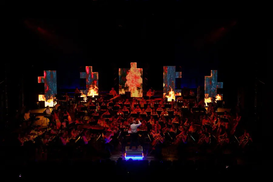 Stesso teatro al buio: orchestra illuminata di rosso davanti a cinque schermi verticali con immagini astratte, direttore al centro
