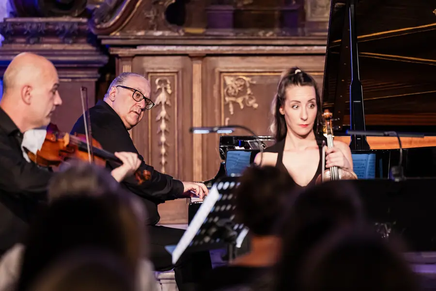 Un uomo con occhiali seduto al pianoforte a coda in una sala con boiserie e stucchi antichi; in primo piano un violinista di spalle e una violoncellista in abito nero che guarda verso il pianoforte