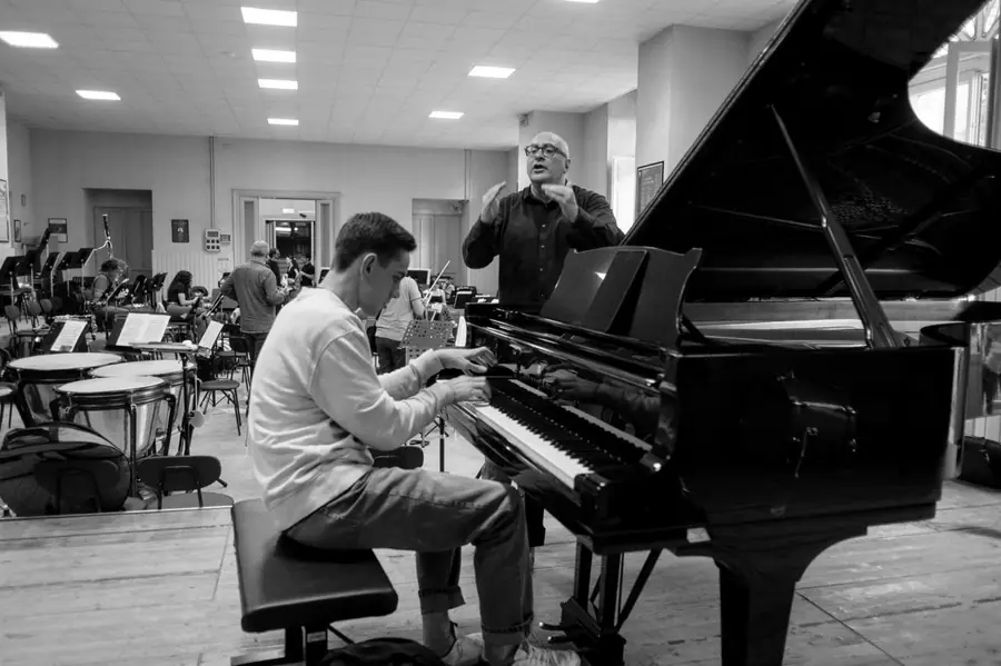 In bianco e nero, entrambe le mani sollevate a modellare una frase musicale mentre il giovane solista suona al pianoforte davanti a lui