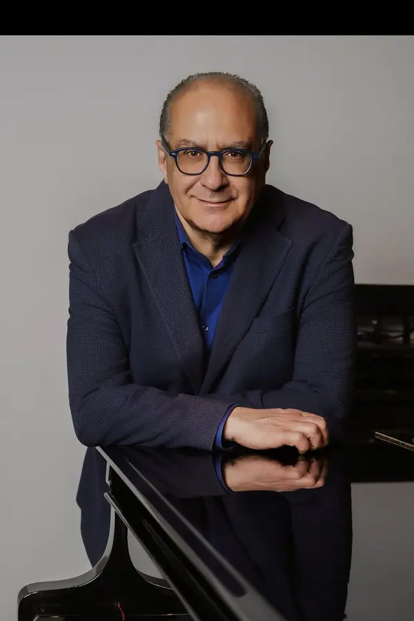 Ritratto verticale in studio: giacca blu e camicia blu, braccia appoggiate al coperchio chiuso di un pianoforte a coda, fondo grigio uniforme
