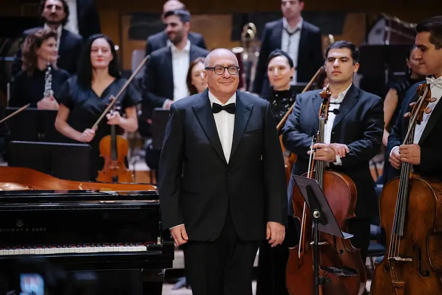 In smoking e papillon, in piedi accanto a un pianoforte a coda Steinway, sorride verso il pubblico mentre l'orchestra applaude alle sue spalle