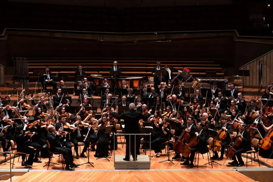 Orchestra sinfonica al completo su un palco di legno chiaro in una sala moderna a terrazze; il direttore e' di spalle sulla pedana, timpani e ottoni in fondo.