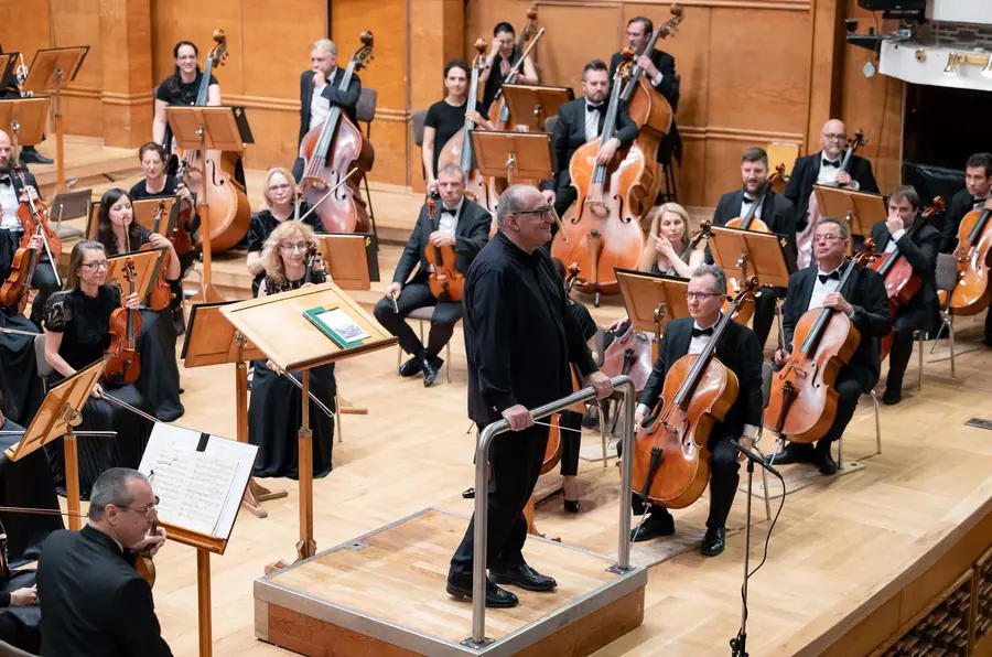Sul podio rialzato al centro della scena, girato verso la sala, con l'orchestra sinfonica al completo disposta intorno in una sala dalle pareti di legno chiaro
