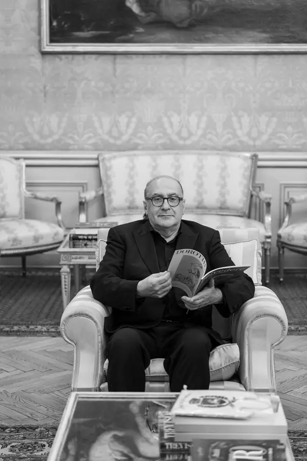 In bianco e nero, seduto in poltrona in un salotto d'epoca con una rivista fra le mani, libri sul tavolino in primo piano
