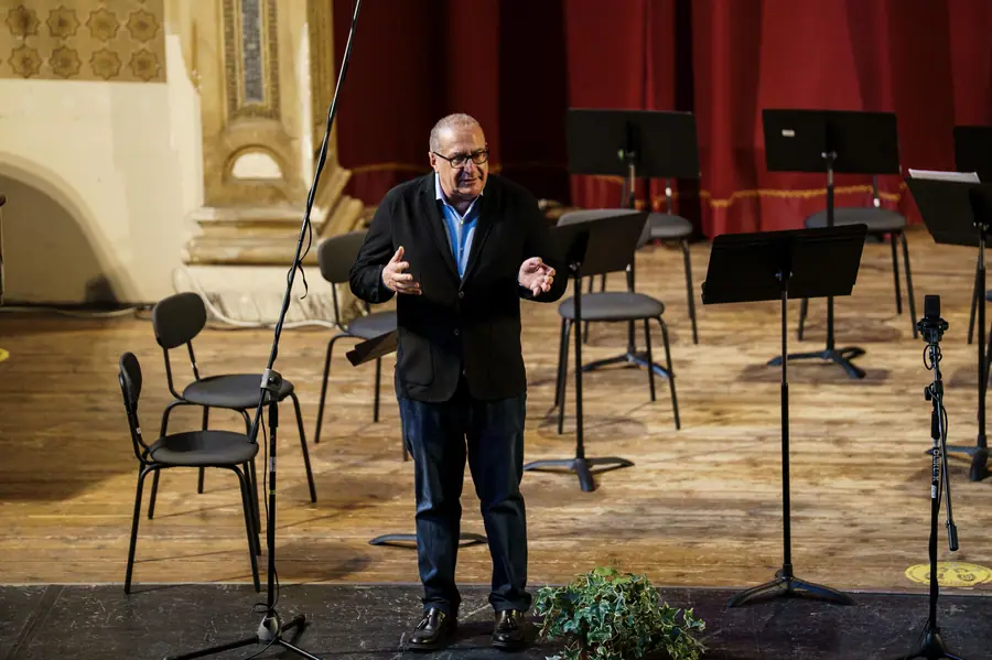 Uomo sui sessant'anni con occhiali, giacca scura e camicia azzurra, parla in piedi sul palco a braccia aperte fra sedie e leggii ancora vuoti, davanti a un sipario rosso.