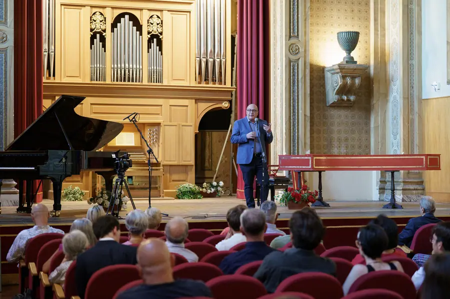 Il palco visto dalla platea: l'uomo in giacca blu al microfono fra il pianoforte a coda, l'organo a canne e il clavicembalo, con le teste del pubblico in primo piano.