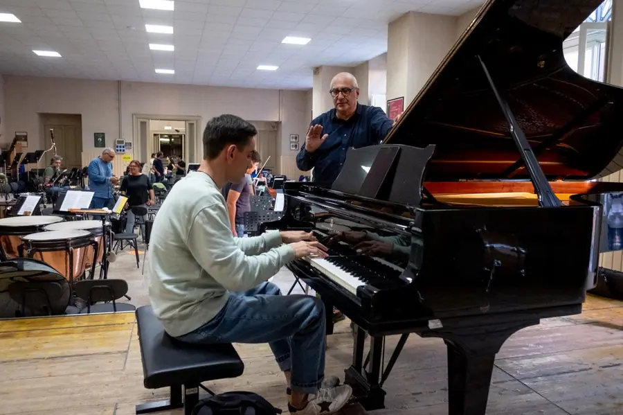 In sala prove alza la mano per indicare un passaggio al giovane pianista seduto al pianoforte a coda, con timpani e orchestra sullo sfondo