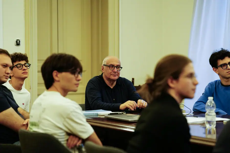 Aula con soffitto alto: l'uomo calvo con occhiali siede al centro dietro un tavolo scuro, circondato da giovani ripresi sfocati in primo piano