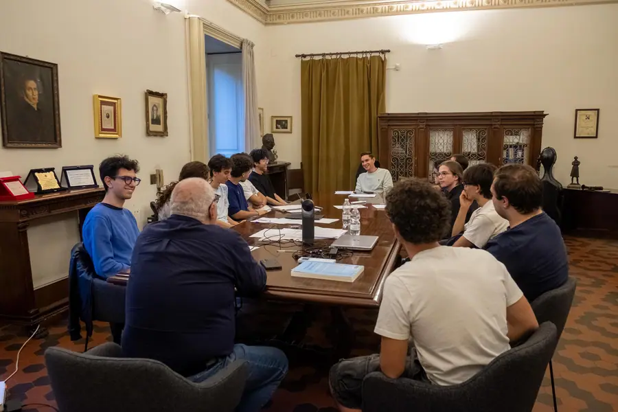 Veduta d'insieme di una sala storica con grande tavolo di legno, ritratti a olio e busti; una dozzina di persone sedute intorno, un uomo anziano di spalle in primo piano
