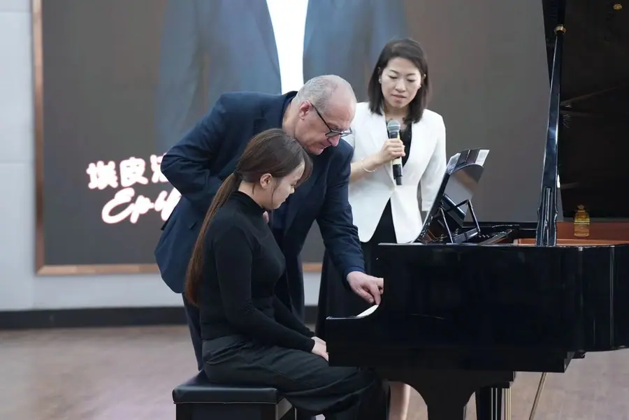 Chino sulla tastiera accanto a una giovane allieva seduta al pianoforte a coda, mentre una donna con il microfono traduce accanto a loro