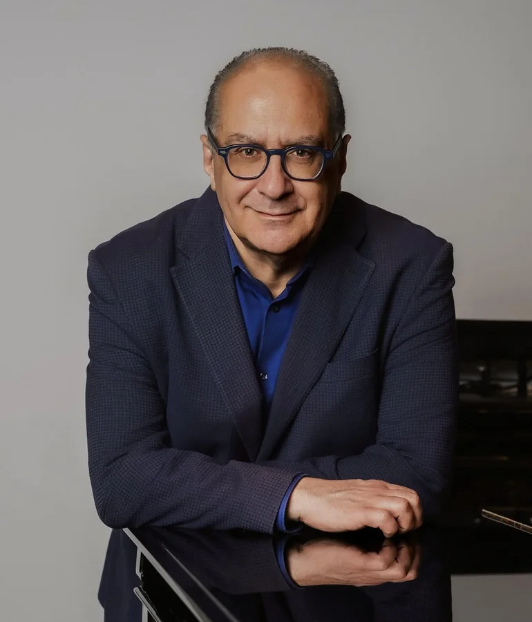 Ritratto a colori: in giacca blu scuro e camicia blu, appoggiato con le braccia al coperchio di un pianoforte a coda nero, fondo grigio chiaro